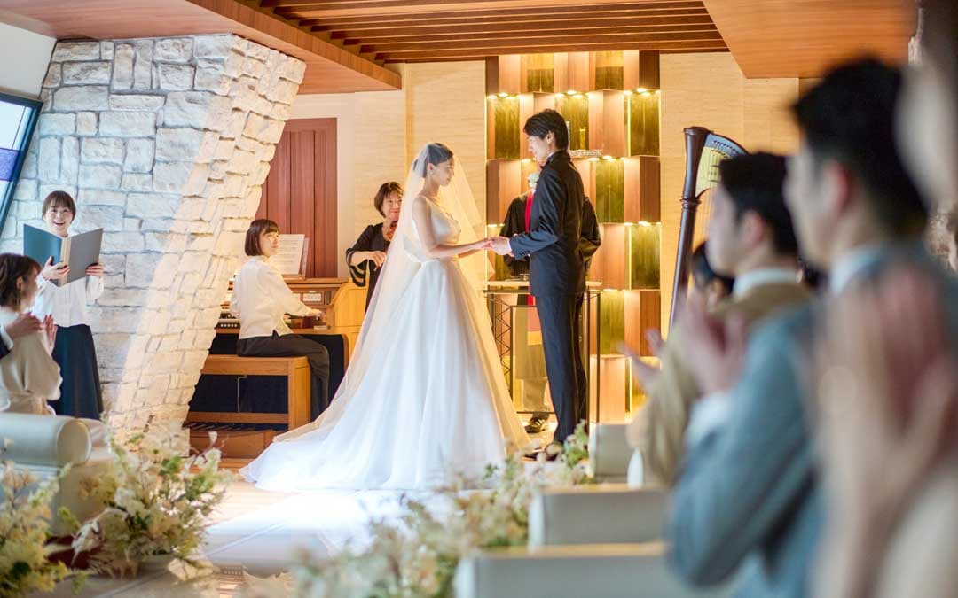 結婚式の儀式中に指輪を交換する新郎新婦の写真。新婦は白いウエディングドレスとベールを身にまとい、新郎はブラックタキシードを着ている。二人は微笑み合い、互いの手を取り合っている。背景には声楽やオルガンを演奏するミュージシャンが見える。