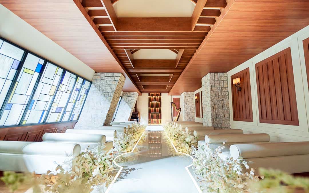 ウエディングチャペル「リガーレ」内観。温かみのある木材と石材を用いた結婚式場。バージンロードは美しく装飾され、左右にはゲスト用座席が配置されている。ステンドグラスの窓から自然光が差し込んでいる。